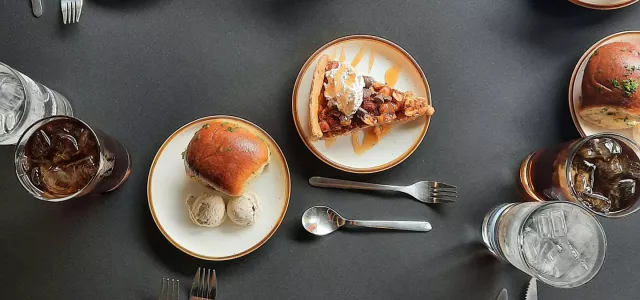 A spread of various foods on plates including as pie, sandwiches, and drinks on a grey table with cutlery sprinkled throughout.