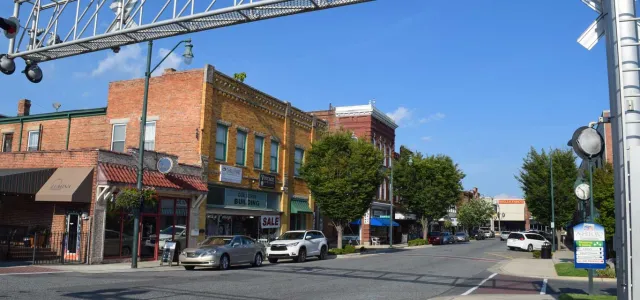 A small town square with a narrow street that is lined with quaint brick buildings and cars parked along the sides. Trees are spaced evenly down the sidewalks under a bright blue sky.