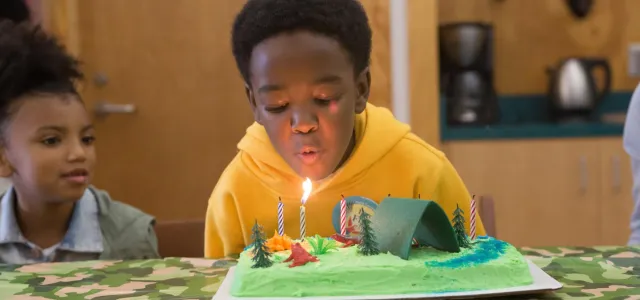 A young child sits at a decorated table, blowing out a candle on a birthday cake. There is another child sitting next to him and a door, sink, and counter with a kettle and a coffee pot on it in the background.