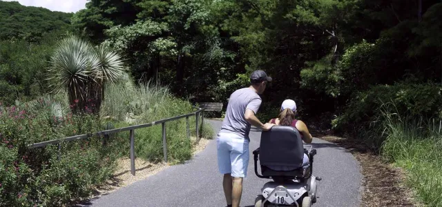 A man wearing a grey shirt and light-colored shorts is standing on a paved pathway, gently guiding or assisting a person seated in a powered wheelchair. The path is surrounded by lush green vegetation, including a unique Yacca plant on the left side, behind a low metal railing.