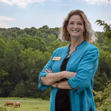 Diane Villa standing in front of a grass field with who rhinos grazing in the background with trees