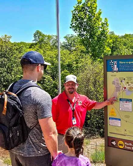 Volunteer standing beside a sign with a large Zoo map talking with a man and his young daugher