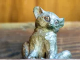 A small, bronze sculpture of a Bush Baby-a small, mousey looking primate with small, rounded ears and large, round eyes sitting in front of a wooden wall.