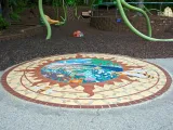 Follow the Pollen Path, a ceramic tile mural, inlaid in front of the Garden Friends playground. This circular mural has an orange sun shape inside with a colorful outdoor scene in the center. 
