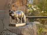 A small bronze statue of an Arctic Fox standing with its signature bushy tail on a rectangular pedestal that has been affixed to the top of a large boulder with trees and a metal handrail behind it.