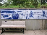  A wide, light wooden bench with a black metal frame sits in front of a long, rectangular mural on a concrete wall. The mural, rendered in shades of blue and white, depicts Arctic wildlife, including walruses and polar bears, and a human figure in winter gear. Green trees and foliage are visible above the wall in the background.