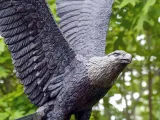 A bronze statue of a Bald Eagle mid-flight, its wings raised and flapping in the wind. A forest of dense trees is visible in the blurred background.