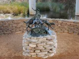 A bronze sculpture of three goats, with swirling forms and intricate details, atop a circular pedestal of stacked, light-colored stones. In the background, a calm body of water and two concrete pillars of a bridge.