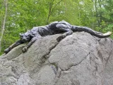 A large bronze statue of a Cougar, a big tan cat with round ears, lying with its body flat and ears back on the top of a giant boulder in a forest.