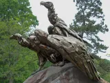 A bronze of three vultures perched close together on a giant boulder, their necks craned and stretched out scanning the wooded area they are in.