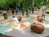 A vibrant outdoor board game with colorful pathways and large brown game pieces shaped like Elephants on a paved circular area. Several children and adults are gathered around and on the game, with lush green trees and foliage in the background, suggesting a park or zoo setting.