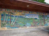 A vibrant, large-scale mural made of square tiles depicts a lush, expansive landscape. On the left, tall trees with light-colored trunks and green foliage frame a scene that transitions into a wide-open field in the center. A light blue river flows from the left foreground to the center. The mural extends across a long wall under a brown overhang.