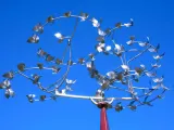 A low-angle shot captures an art installation that is designed to spin in the wind like a weather vane against a clear, vibrant blue sky. It is made of several spiraling, circular, silver tubes that are lined with smaller, silver shapes that look like wings. As the piece spins, it looks like butterflies fluttering in a circle. The central support structure is a reddish-brown pole, visible at the bottom of the sculpture. 