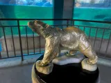 A small bronze statue of a Polar Bear walking on ice that has been placed on top of a pillar. There is a metal handrail lining a ramp that leads to a glass viewing area of an animal habitat pool in the background.