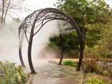 An intricate, domed archway with a tree branch design, that spans a sidewalk that is lined with a variety of plants. There is water being sprayed down in a fine mist to keep those who walk underneath it cool.