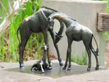 A small bronze statue of two Giraffes standing in front of each other, their necks bending down and crossing each other to look at a baby Giraffe that is laying between them. This artwork is affixed to the top of a large stone.