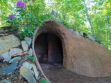 A whimsical, hobbit-hole-like earthen structure with a rounded archway entrance is nestled into a hillside in a lush green forest, blending seamlessly with the surroundings. To the left of the entrance, a stone wall is integrated into the structure, and a vibrant purple rhododendron bush with green leaves blossoms atop the earthen roof.