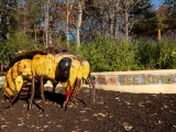 A gigantic statue of a worker bee stands on the ground at the Garden Friends Playground with its wings tucked back. A mosaic wall stands behind it with tiled words that read "Bee Kind" and various small flowers.