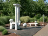 Zoological egg rest art installation which is comprised of minimalist white stone sculptures in a lush green outdoor setting, with a tall central piece, spherical elements, and a bench-like form beside a paved walkway.