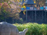 A lifesized statue of a Hippopotamus mom and calf standing on a mulch covered shore of a lake, tall reeds visible in the background. In the distance, a wooden pier with colorful handrails and a round rotunda spans the water.