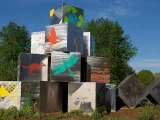 A striking silver cube sculpture, representing themes of conservation and nature, stands on a grassy mound near a pathway. The cubes are adorned with brightly colored environmental and wildlife graphics.
