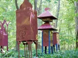 A group of tall, artistic metal sculptures resembling red-brown lanterns or towers, featuring animal cut-outs, are displayed outdoors in a dense wooded area.