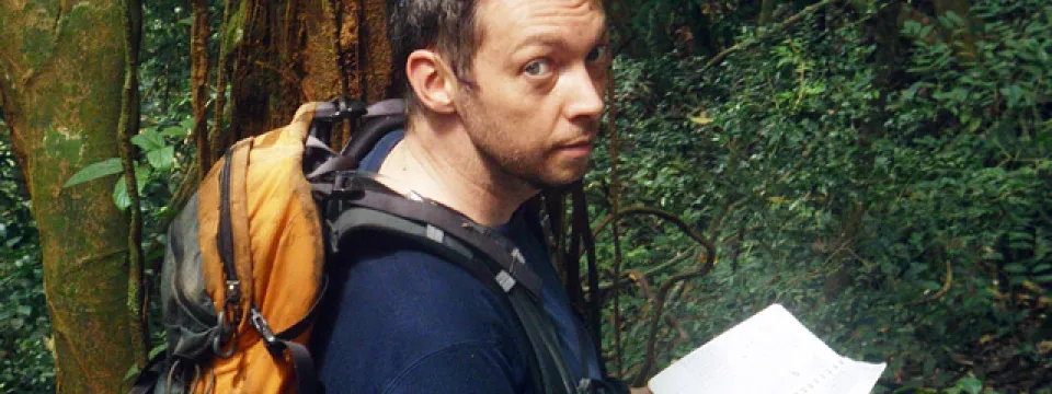 A man with short brown hair wearing a navy blue shirt and an orange backpack looks over his shoulder at the camera. He is holding a paper and pen and is standing in an African rainforest, surrounded by thick green trees and underbrush.