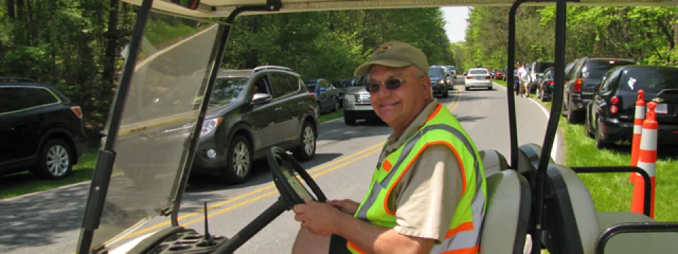 An older man driving a white golf cart on a road with cars parked on the side. He is wearing a neon vest and a hat.