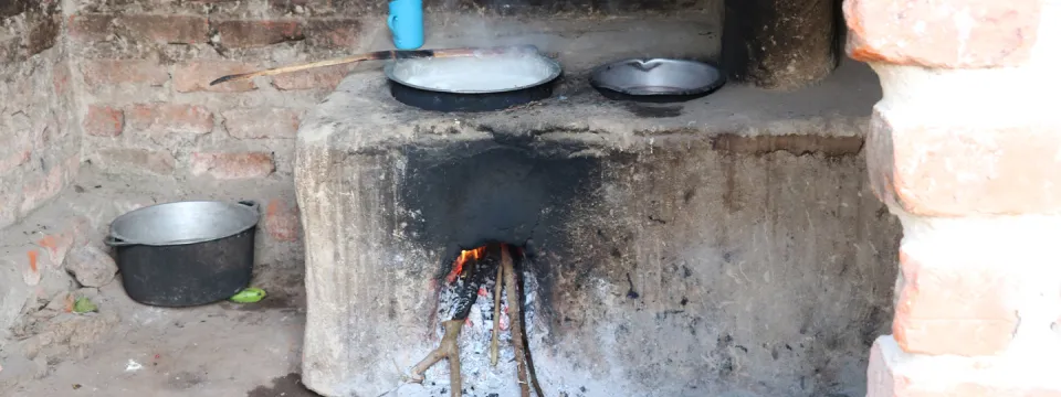 A traditional earthen cookstove, is shown with two pots on top. Flames are visible inside the firebox. A blue mug and a metal bucket are placed nearby. The cookstove is constructed from mud and bricks and appears to be inside a brick room.