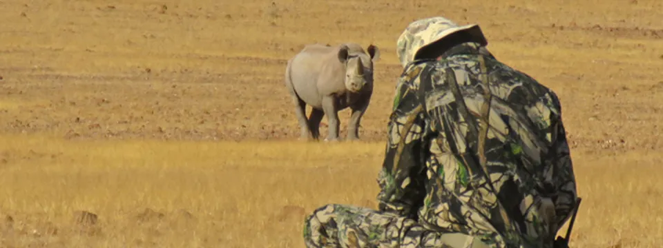 A person wearing camouflaged colored clothes crouched down conducting a rhino monitoring with Save the Rhino Trust. There is a rhinoceros in the background facing the person in a savanna environment.