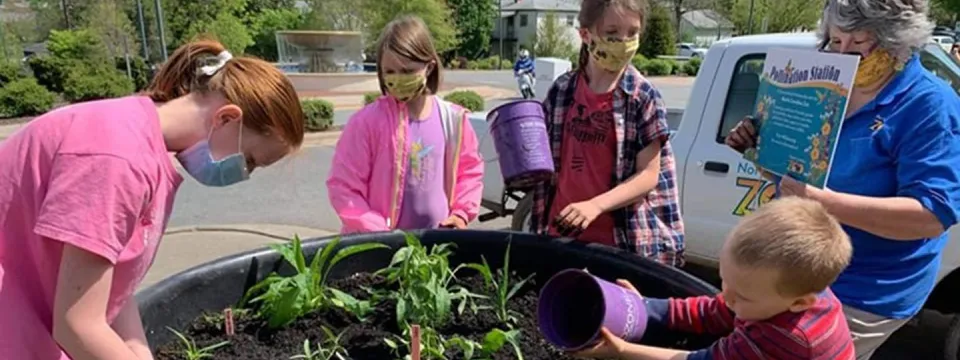 Four children wearing face masks and an adult wearing a face mask are gathered around a large, black planter filled with plants. One child is pouring water from a purple container into the planter, while another holds a similar container. The adult is holding a small sign that says "Pollination Station." A white truck and a parking lot are visible in the background.