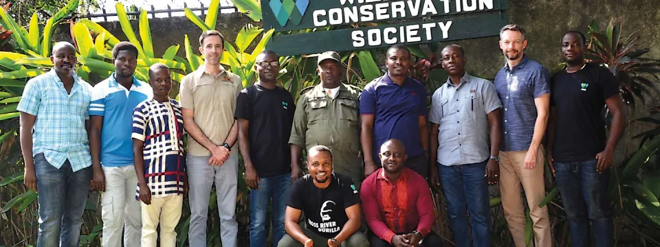 A group of eleven men are posing for a photo in front of a sign that reads "WILDLIFE CONSERVATION SOCIETY". The group appears to be diverse in terms of perceived race and ethnicity. Most of the men are standing, while two are kneeling in the front. They are dressed in a variety of casual clothing. Behind them, there is a wall or fence with lush green vegetation. 