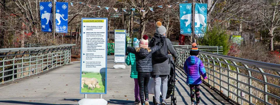 A group of Winter guests entering the Zoo. Three individuals including one adult and two kids walk over a large cement bridge that has tall handrails on either side and two tall informational signs placed at either end. In the background are trees, colorful pennant flags, and blue banners with pictures of Winter animals on them.