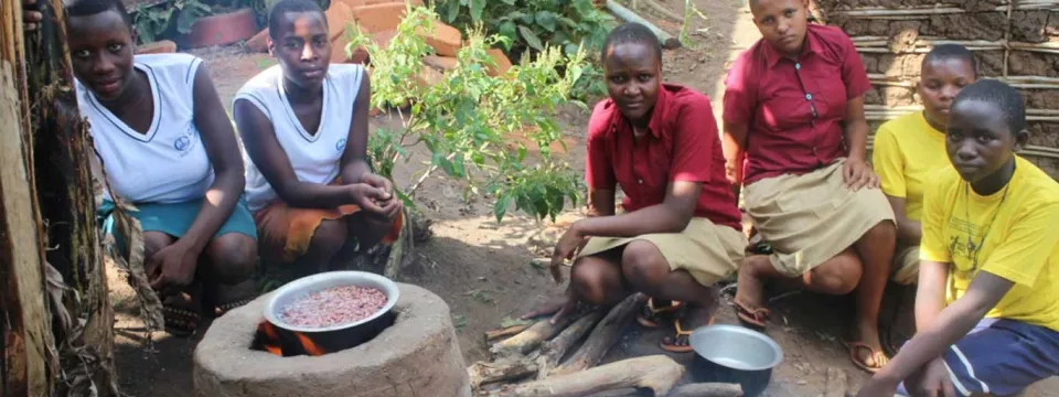 Six young women or girls are crouching around a traditional outdoor cooking fire set up on the ground, likely in a rural African community. In the center, a cooking pot filled with red beans sits on a raised clay or stone stove. The girls are looking towards the viewer, wearing a mix of school uniforms (white vests and skirts/wraps, or maroon shirts) and yellow t-shirts. More cooking pots and wood are visible on the ground.