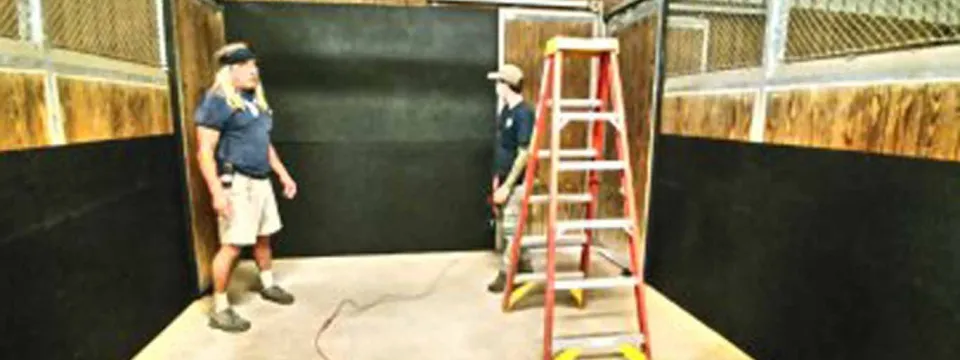 Two zoo staff members set up a large black mat on the floor of an empty interior animal stall, using a red ladder nearby.