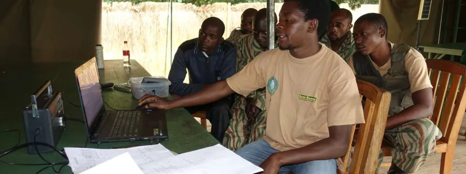  A man wearing a tan t-shirt with a green logo is sitting at a table inside a canvas tent, working on a laptop computer, gesturing towards the screen and appears to be leading a session or training. Several men are seated behind him, listening and watching, wearing clothes that suggest a uniform or ranger attire. Papers are spread out on the table in the foreground, and radio equipment is visible on the left.