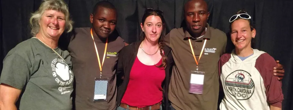 A diverse group of five people stand together, arm in arm, posing for a photo against a black background.