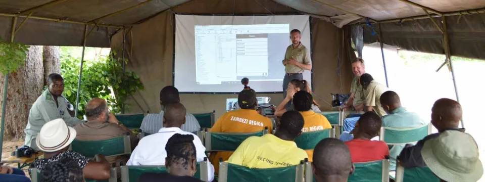 A small group of people is attending an outdoor presentation held under a large tent. A man in a light-colored shirt is standing in front of a projection screen displaying a data table. The audience is seated in rows of chairs, facing the screen. Several people in the audience are wearing yellow shirts that have writing on the back (partially visible). The surroundings outside the tent appear green and leafy.