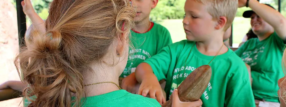 A group of young children are gathered closely, engaged in an activity that involves handling a long, reddish-brown, smooth object that looks like a rock or a tool. The focus is on a girl in the foreground, seen from the back, with her long blonde hair in a ponytail, and a boy in a green shirt facing her, touching the object with his hand. Another boy in a green shirt is visible behind them, and an adult's head is partially seen in the background.