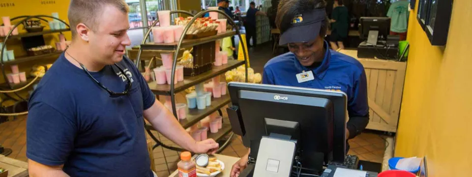 A restaurant worker stands at a cash register ringing up the food purchases of another person who is standing on the other side of the counter. A display of cotton candy stands behind them on a circular rack.