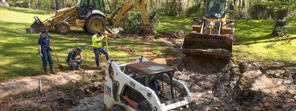 An outdoor construction scene showing three pieces of heavy machinery and three workers involved in digging a trench in a grassy, wooded area. In the foreground, a white skid-steer loader is positioned in a muddy trench. In the background, a yellow backhoe and a yellow front-end loader are also working. Two workers are standing or kneeling near the trench, and one worker is operating the skid-steer loader.