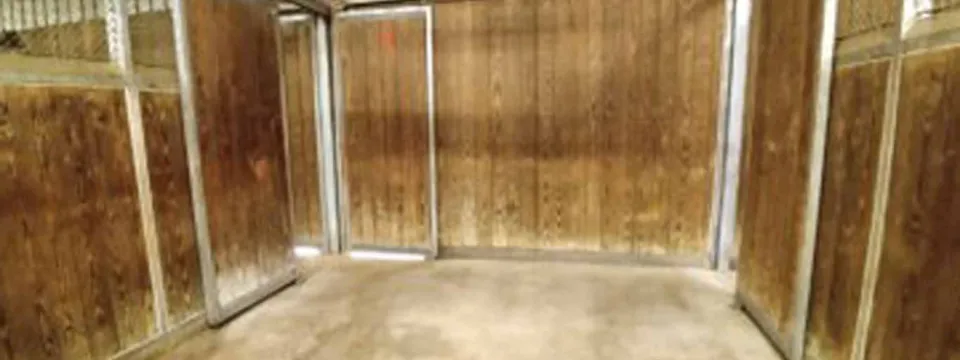 An empty horse stall with three sides made of wooden planks and a metal frame. The floor is concrete, and the stall is located inside a large barn-like building with a metal ceiling.