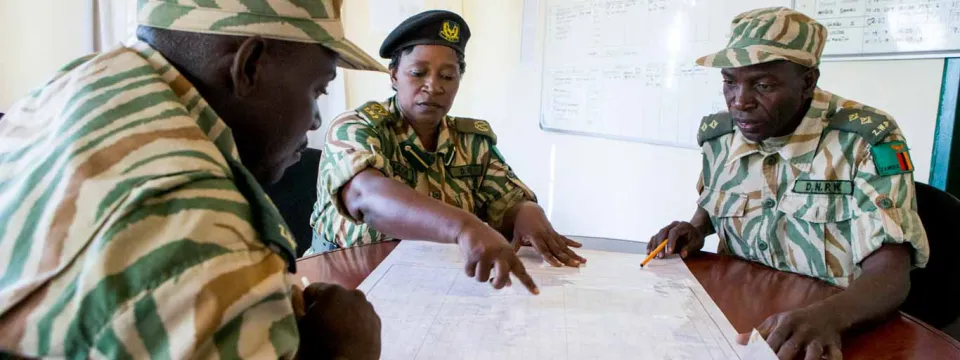 A group of three park rangers in tan and green uniforms sit around a round wooden table looking at and pointing to a point on a map that lays between them.