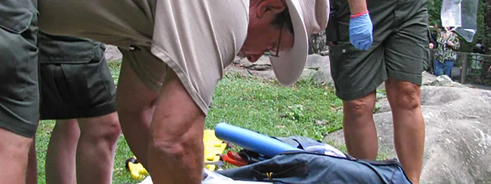 A park ranger or field medic, wearing a wide-brimmed hat, tan shirt, and blue gloves, bent over an open orange and black medical or field kit. The ranger is surrounded by other people in similar uniforms, standing on rocky ground with grass and foliage in the background, suggesting an emergency response or first aid situation.