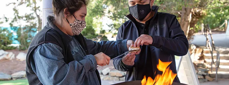 A man and a woman wearing Fall attire and face masks, stand side by side next to a fire pit, making S'mores. They appear to be standing under a covered patio with a large white column, and trees visible behind them.
