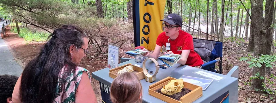 Young adult volunteer at the Investigation station talking to a mother and two children