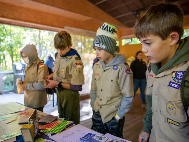 Four young people appear to be gathered around a table outdoors. They are wearing tan boy scout uniforms that are covered with patches and appear to be listening to another individual who is not visible to the viewer. On the table are various objects, including what appear to be drawings and graphs. The setting looks like a covered outdoor area with stone pillars or walls in the background.