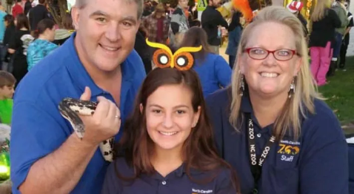 Three smiling community educators, one male and two female, holding various animal ambassadors, including a snake, tarantula, and two hissing cockroaches, at a Fall Festival event with crowds of eventgoers in the background.