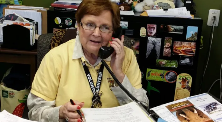 An older woman with glasses and short, red hair, wearing a yellow collared shirt, appears to be in an office talking on a corded telephone while sitting at a desk that is covered with papers. There is a black filing cabinet covered with magnets behind her.