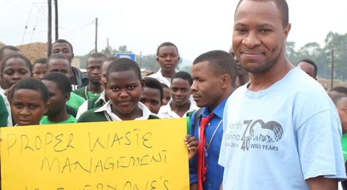 Bruce, a young, dark-skinned man wearing a blue NC Zoo shirt smiles as he stands with a group of dark skinned children of varying ages from with Bigodi secondary school club. Two of the children hold a yellow sign with the words, "Proper waste management is everyone's responsibility" written in black marker.